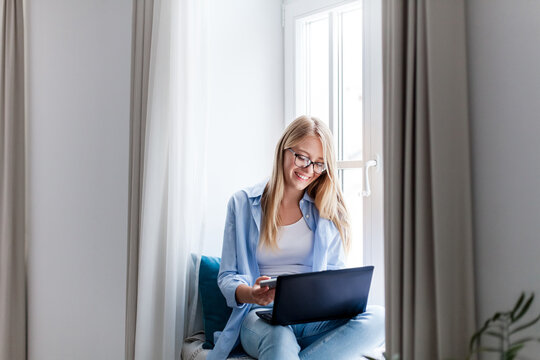 Happy woman working from home office. Smiling freelancer using laptop and mobile phone. Cozy workplace in living room on windowsill. Concept of female business, career, teleworking, wellbeing