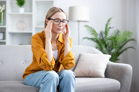 Young woman feeling intense head pressure, touching temples with closed eyes, experiencing stress, exhaustion, or a migraine while sitting on a sofa at home