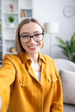 Young adult woman with short blonde hair and eyeglasses holding a smartphone, capturing a cheerful selfie, standing in a bright home interior while looking directly at the camera