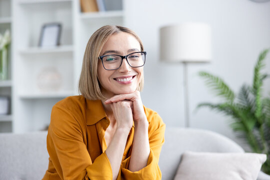 Blonde woman with bob and eyeglasses smiling confidently on a comfortable sofa in a bright modern home office, smart-casual and relaxed during remote work or a video call