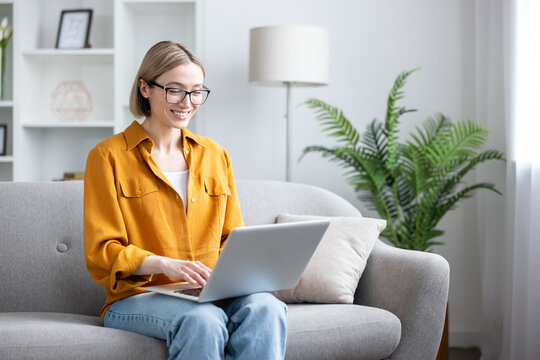 Happy young woman in glasses smiles while typing on a laptop, teleworking from a cozy sofa in her bright home office, relaxed, focused and comfortable with remote work