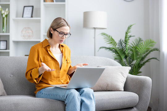 Young woman sitting on a couch at home, working remotely on her laptop, expressing confusion and frustration with an issue or problem encountered while shrugging and looking at the screen