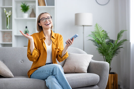 Young woman wearing glasses laughing with joy while sitting on a comfortable couch in a bright living room, holding a smartphone and reacting to good news, success, or a funny message