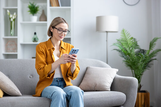 Young woman wearing glasses sitting comfortably on a sofa in a modern living room, smiling while engaging with her smartphone, representing connectivity, leisure, and digital communication