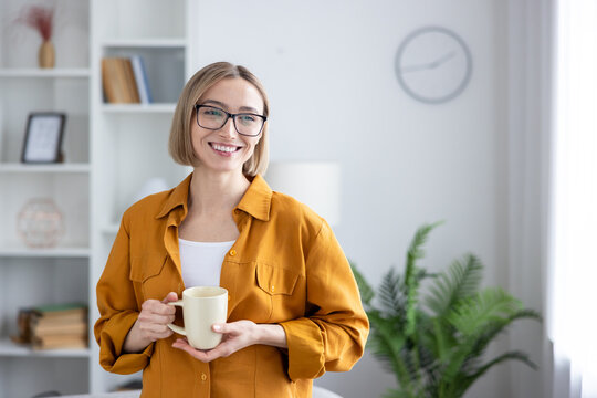 Young woman with glasses smiling, enjoying a warm mug during a relaxed break in her bright home office, radiating comfort, confidence and cozy, productive vibes
