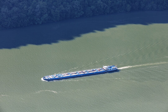 Aerial top-down view of a blue inland tanker barge navigating a wide green river, leaving a wake. Freight transport concept.