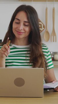 Vertical HD video of young creative woman thinking and writing down ideas in a notebook while working on her laptop from a modern kitchen, showcasing a productive remote work or e-learning lifestyle