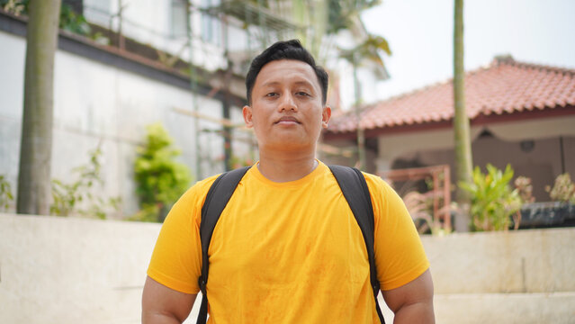 Portrait of a confident young Asian man in a yellow shirt wearing a backpack, standing outdoors looking at the camera. Casual lifestyle concept of a student or traveler in a tropical neighborhood.