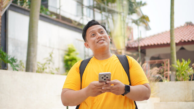 Young Asian man in a yellow shirt holding a smartphone with both hands, smiling and looking up thoughtfully outdoors. Casual lifestyle concept of a student or traveler using mobile technology.