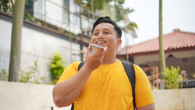 Person in a yellow shirt using an AI voice assistant on a smartphone, speaking a voice command to the glowing orb on screen showing "what can I help you with?" Modern mobile technology concept.