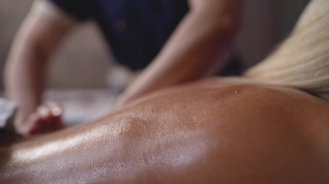 Soft focused closeup of woman's oiled back during massage. Warm skin tones, gentle lighting, blurred therapist hands in background. Evokes calm, luxury, wellness