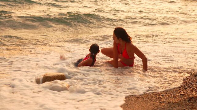 Family vacation enjoying sea surf with smiling mother and daughter lying down on pebbled beach shore. Static shot.