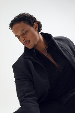 Profile image of a fashion studio shot of an African American man wearing a black coat, posing against a white background, looking at the down.