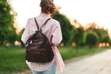 Fototapeta premium Woman with backpack walking along a sunlit park path, casual outfit, warm breeze, vibrant greenery, serene outdoor moment