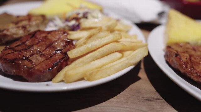 Grilled pork steak served on a white plate with crispy golden french fries, savory gravy sauce, and a fresh side salad. Close-up view of a delicious restaurant meal under warm ambient lighting.