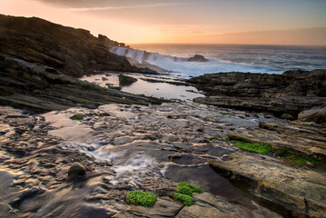Fototapeta premium Sandstone rock formations with tafoni erosion on the coast of Jaizkibel during a dramatic sunset with long exposure ocean waves flowing over rocks in Jaizkibel, Basque Country, Spain
