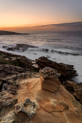 Fototapeta premium Sandstone rock formations with tafoni erosion on the coast of Jaizkibel during a dramatic sunset with long exposure ocean waves flowing over rocks in Jaizkibel, Basque Country, Spain
