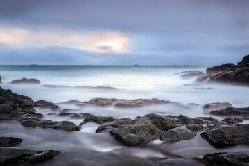Fototapeta premium Dramatic pink sunset sky with long exposure ocean waves washing over a dark pebble beach and coastal hills in Ogeia Beach