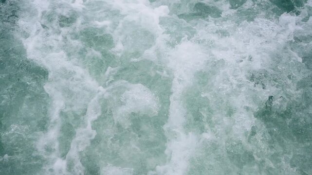 Close up top view of turbulent blue ocean water with white foam and bubbles. Powerful churning sea waves creating a textured splashing pattern. Nature background of rushing river or boat wake.