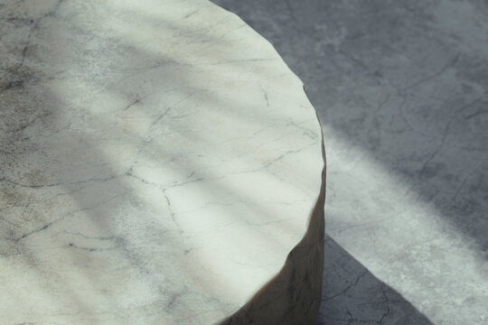 Minimalist 3D render of round beige marble table with a rough raw edge on a concrete floor in soft natural sunlight. Empty display stage with botanical window gobo shadow and copy space for cosmetics.