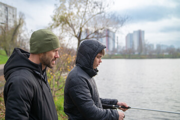 Fototapeta premium Father and teenage son fishing together on a lake shore in an urban environment with city skyline