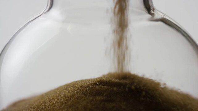 Slow motion static zoom handheld shot of sand grains pouring and accumulating into glass jar hourglass on white studio background