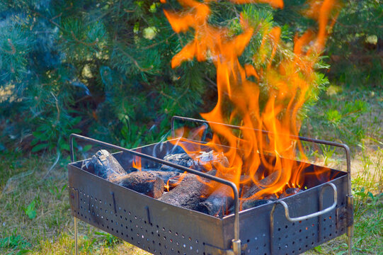 Intense orange flames burning in a metal charcoal grill outdoors with green pine trees in the background on a sunny day