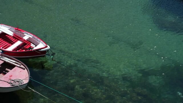 High angle view of two small red and white rowing boats moored in crystal clear, turquoise-green sea water. Sunlight sparkles on the gentle ripples, revealing the rocky seabed and mooring chains bene