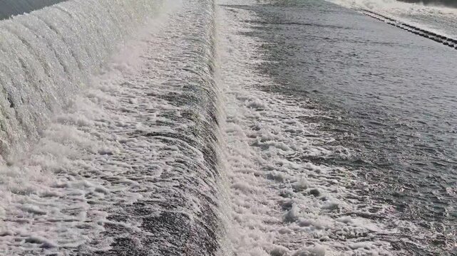 Medium shot showing river water cascading over an inflatable rubber dam crest, creating continuous whitewater, splashes, and turbulent hydraulic current down the sloping barrier face.
