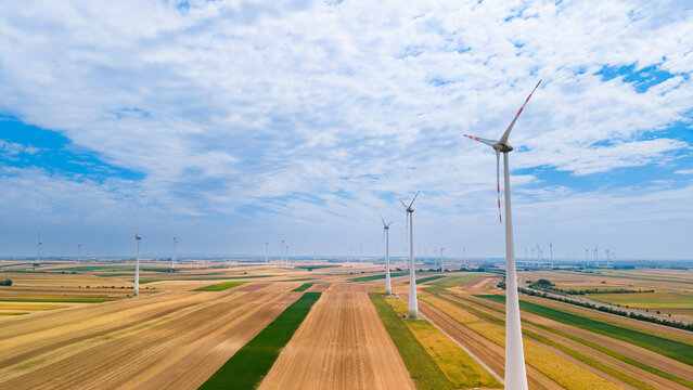 Wind turbines stand tall in fields under a blue sky with clouds. Wind turbines spin in open fields during a sunny day with few clouds. The landscape shows various crop patterns.