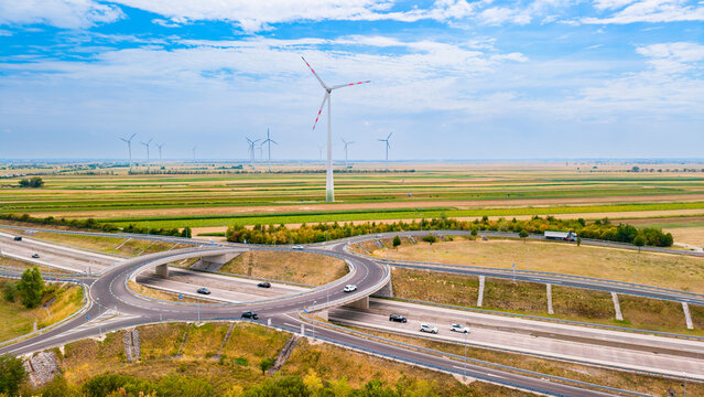 Wind turbines stand tall near a highway with passing cars on a sunny day. Vehicles drive on a roundabout located beside wind turbines and fields under a blue sky.