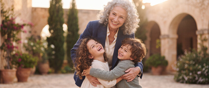 Happy grandmother hugging her smiling grandchildren outdoors in a sunlit Mediterranean villa courtyard, capturing genuine family love, intergenerational joy, and togetherness