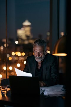Serious attorney reviewing contracts late at night in a modern office, illuminated by laptop light with city lights visible in the background.