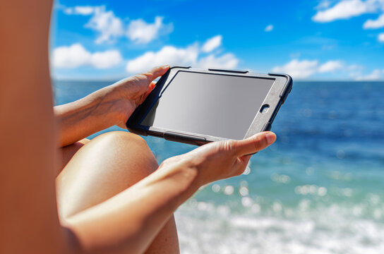 A person holds a digital tablet with a blank screen against a beautiful ocean backdrop on a sunny day.