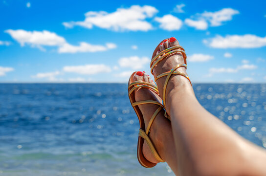 A woman relaxes on a tropical beach with her feet up in gold sandals against a bright blue ocean and sky.