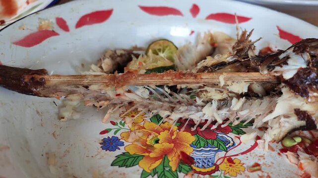 Close up view of a fully eaten fish skeleton and spine bone skewered on bamboo lying on a dirty vintage enamel plate after lunch at a restaurant.
