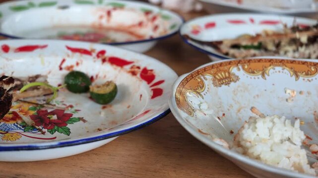 Wide panning shot showing multiple dirty plates, bowls with leftover rice, fish bones, and empty glasses scattered on a restaurant table waiting to be cleaned.
