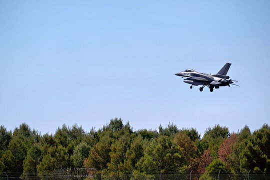 A military jet approaches for landing just above a dense treeline, descending against a clear blue sky. This scene captures dynamic aviation moments for enthusiasts.