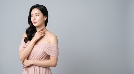 Obraz premium Elegant young asian woman with long wavy hair wearing a soft pink offshoulder dress, posing gracefully against a clean light gray studio background