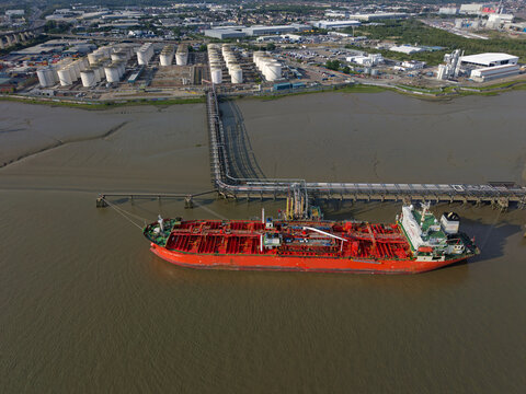 Aerial view of red oil tanker moored by oil terminal near Dartford Crossing in UK. Oil tanker on river Thames delivering fuel. Pipeline leading towards white fuel storage units.