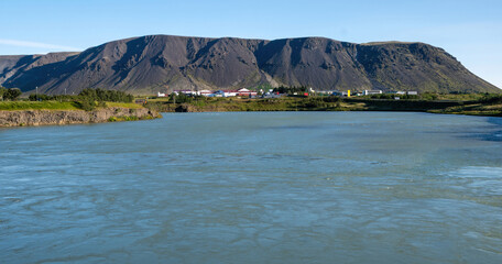 Naklejka premium A scenic view of a wide river flowing through Selfoss with a dramatic volcanic mountain and a small town in the background under a clear blue sky.