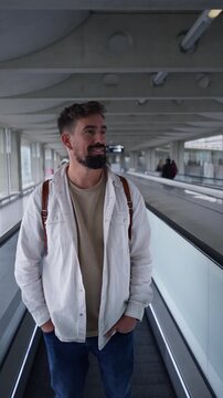 Vertical video of a bearded caucasian traveler standing with hands in pockets on a moving walkway, calmly riding through a spacious airport terminal.