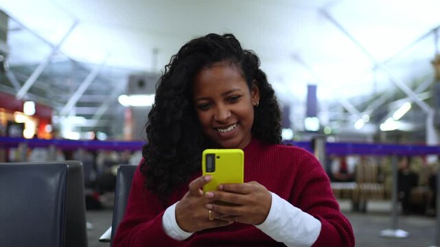 An upbeat styled shot of a woman in a red knit sweater sitting in a bright airport lounge, focused on a yellow smartphone, capturing casual travel moment and informal communication.