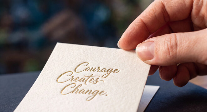 Closeup of a hand holding a textured card with the gold embossed text courage creates change on a dark background