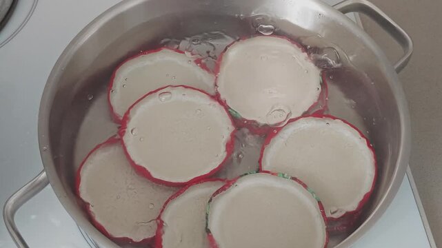 Close up view of sterilizing glass jar lids in a steel pot of boiling water. Home canning preparation for food preservation on a hob