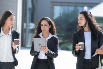 Three female colleagues in matching black business suits, talking as they walk in step, move as a group across a sunlit forecourt with a white tote bag and printed papers. © ultramansk