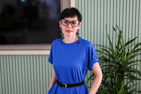 Portrait of a beautiful brunette woman wearing glasses and an elegant blue dress, smiling confidently while looking at the camera indoors.