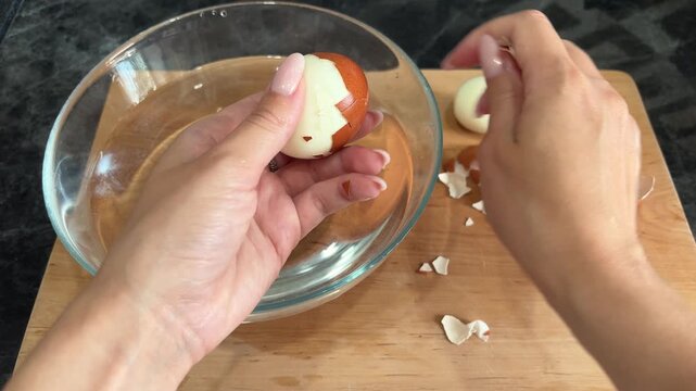 Woman peeling a boiled egg by hand over a bowl, simple home cooking, kitchen preparation, healthy protein food, everyday meal routine.