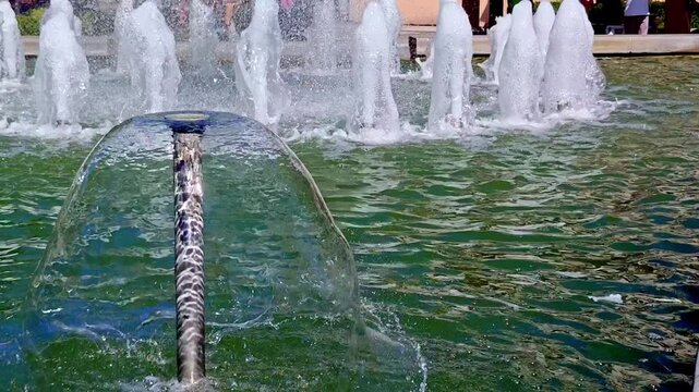 Close-up of a fountain nozzle creating a smooth glass-like dome of water in a public city fountain, with multiple spouting foam jets in the background and rippled greenish water surface in daylight.