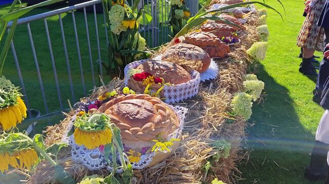 Row of traditional, ornately decorated harvest festival bread loaves arranged on straw bales, adorned with fresh sunflowers, lace napkins, and flowers outdoors on a green lawn in daylight.
